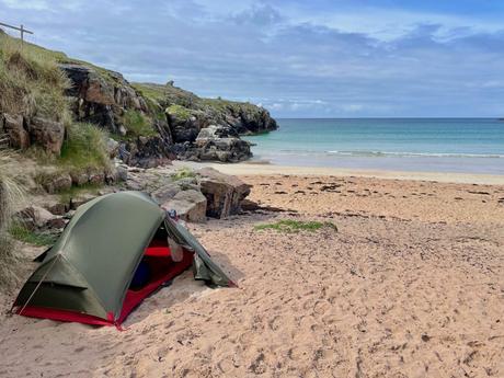 A green camping tent pitched on a sandy beach near rocky cliffs, overlooking calm, crystal-clear waters on a bright day.