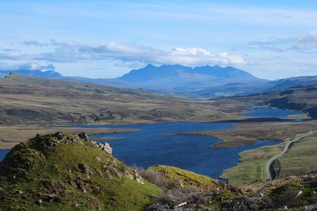 A scenic valley with a large, blue lake stretching into the distance, surrounded by rolling green hills and towering mountain ranges under a clear sky.