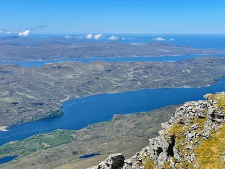 An aerial view of a deep blue loch surrounded by rolling hills and rugged terrain, stretching toward the horizon under a clear blue sky with a few scattered clouds.