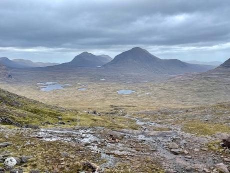 A wide, expansive view of a valley floor in the Wester Ross area of the Northwest Highlands, dotted with small lakes, surrounded by towering mountains under a moody, overcast sky.