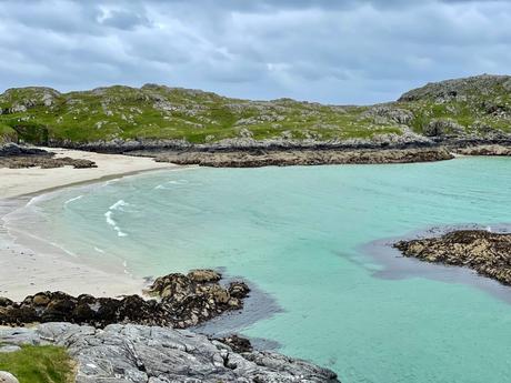 A scenic cove with turquoise water gently lapping onto a white sandy beach, surrounded by rocky outcrops and lush green hills.