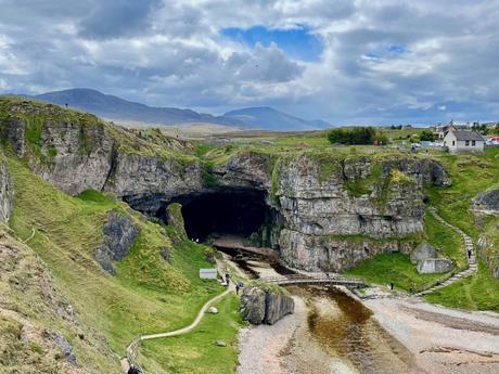 The large mouth of Smoo Cave set into a steep cliffside, surrounded by grassy slopes and pathways, with visitors exploring the area below.