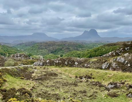 A moody landscape featuring rolling green hills, with distant jagged mountains standing under a cloudy sky.