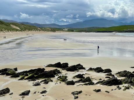 A wide, expansive beach with wet sand reflecting the sky, scattered rocks in the foreground, and distant hills in the background.