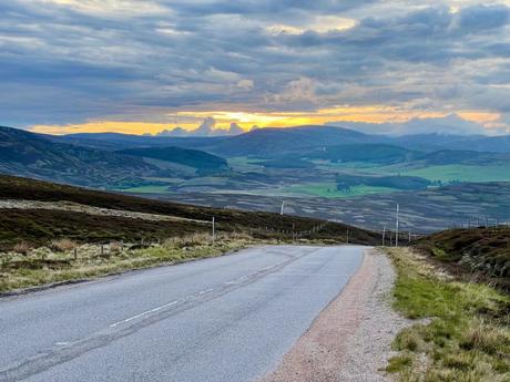 A long, winding road cutting through a hilly landscape, with the sky painted in warm hues of a setting sun, casting soft light on the expansive valleys below.