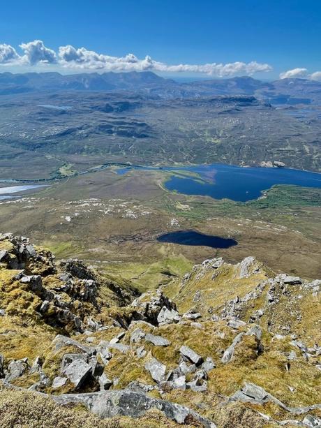 A vast landscape seen from the top of Ben Hope, showcasing a mixture of rocky outcrops, patches of green, and small lakes scattered across the valley below, with distant mountains on the horizon.