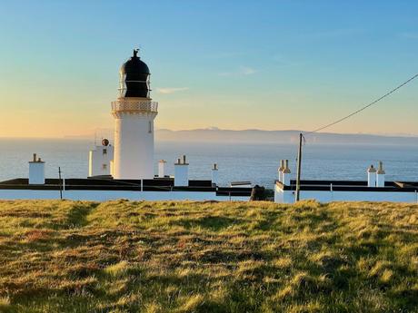 A picturesque lighthouse standing proudly on a grassy clifftop at sunset, with the ocean stretching out behind it, and the Orkney Islands in the far distance, all bathed in warm golden light.