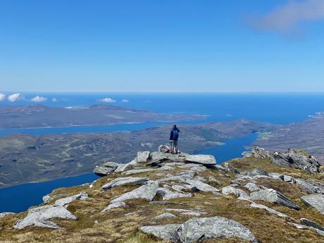 Alex Tiffany standing at the edge of a rocky peak at the summit of Ben Hope, gazing out over a stunning landscape of lochs, rolling hills, and distant islands, all beneath a clear blue sky. The expansive view captures the tranquil beauty of the Scottish Highlands, with water and land stretching into the horizon.