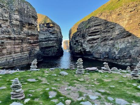 A striking coastal scene featuring tall, layered cliffs that rise dramatically from the water, with several rock cairns built by visitors scattered along the grassy foreground.