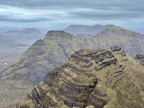 A panoramic view of rugged mountains with steep ridges under a cloudy sky, the rocky terrain creating dramatic contours across the landscape.