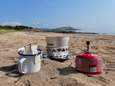 A portable coffee brewing setup with a mug and a Primus stove, placed on a sandy beach with a scenic coastal view in the background.