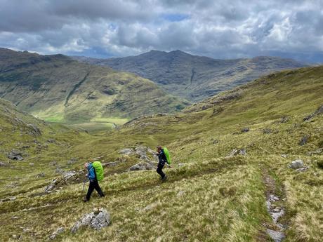 Two hikers ascending a grassy hill, surrounded by vast, green valleys and towering mountains under a partly cloudy sky.