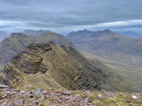 A panoramic view of a rugged mountain landscape, with steep ridges and distant peaks, under overcast skies.