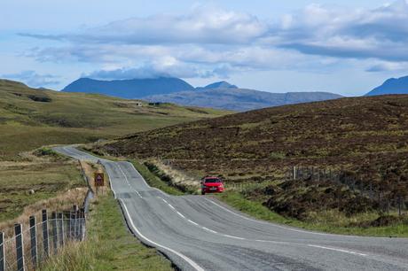 A winding rural road on the Isle of Skye leading into a hilly landscape, with a red car parked by the side of the road and mountains in the background under a cloudy sky.