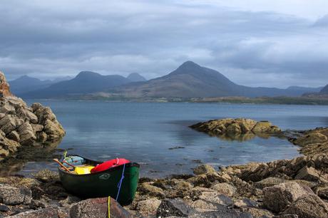 A green canoe resting on a rocky shore beside a calm lake, with towering mountains in the distance under a cloudy sky.