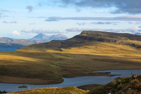 A landscape of open moorland and gently sloping hills, with sunlight casting golden hues on the landscape and distant mountains under a partly cloudy sky.