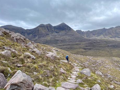 A lone hiker making their way up a rocky path surrounded by steep, grassy mountain slopes and imposing peaks in the distance.