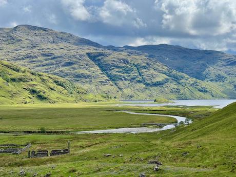 A wide shot of a lush, green valley with a river curving through it. In the foreground, the ruins of a small stone building stand, while the mountains in the distance loom under a partly cloudy sky.