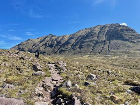 Two hikers walking along a rocky trail towards a large mountain, under a clear blue sky, with vast open landscapes around them.