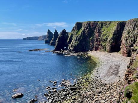 The rugged Scottish coastline at Duncansby Head with towering sea stacks and steep cliffs, with the blue sea gently lapping at the rocky shoreline.