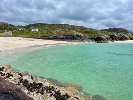 A serene beach with white sand and emerald water, bordered by rugged rocky hills, with a few cottages dotting the landscape.