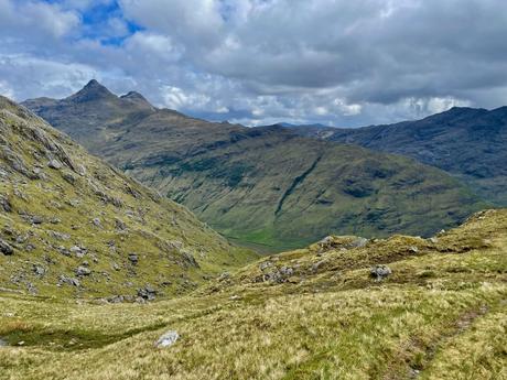 A rocky path leading through a mountainous valley, with jagged peaks on both sides and patches of green grass along the trail.