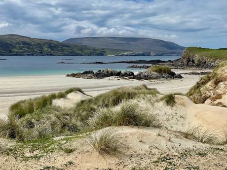 A tranquil beach scene with pale sand dunes covered in grass, leading down to calm blue waters and rocky outcrops in the distance.