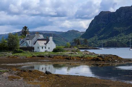 A white cottage in Plockton with chimneys, nestled near the shore of a bay, surrounded by green hills and distant mountains, with sailboats floating on the water.