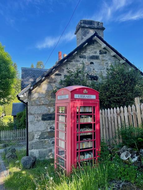 A red phone box converted into a small public library stands in front of a quaint stone house, surrounded by greenery and under a clear blue sky.