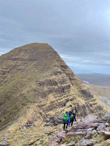 Two hikers with backpacks walking along a mountain ridge, descending a rocky trail surrounded by vast, barren terrain under a cloudy sky.