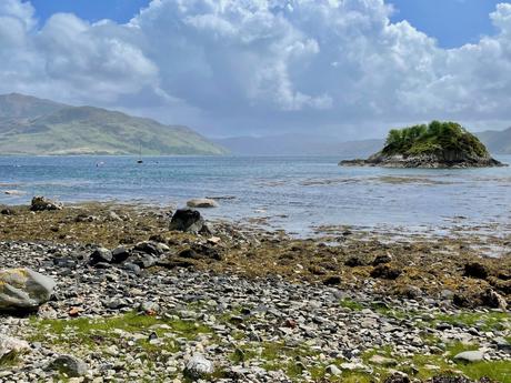 A scenic coastal view featuring a rocky shoreline with patches of green moss. The calm sea stretches out to a small, rocky island topped with a few trees, while the distant hills are shrouded in clouds under a bright, partly cloudy sky.