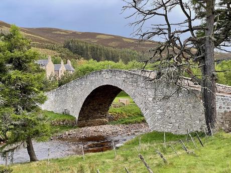 A charming old stone bridge arching over a shallow stream, surrounded by trees and cottages nestled in the Scottish countryside.