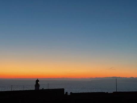 A stunning sunset over the ocean with a silhouette of a lighthouse in the distance, the sky transitioning from deep orange to dusky blue.