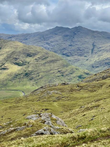 A rugged mountain landscape in Knoydart with steep, rocky slopes and a muted green covering. The sky is overcast, casting a grey tone over the scene with distant peaks visible in the background.