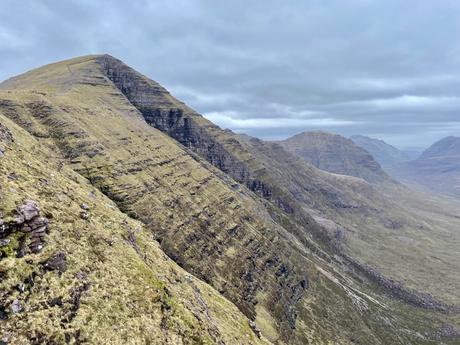 A steep, rocky mountain slope stretching into the distance, with layers of ridges and valleys under overcast skies.