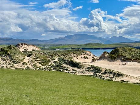 A lush green field with sand dunes in the background, bordered by turquoise waters, with mountains rising in the far distance under a partly cloudy sky.