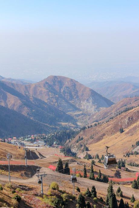 Multiple gondola lifts travel up a dry, rolling mountain valley with scattered trees, buildings, and steep slopes, under clear skies.