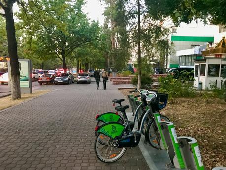 A street scene with a row of bicycles lined up at a docking station on a tree-lined sidewalk. In the background, parked cars and pedestrians are visible.