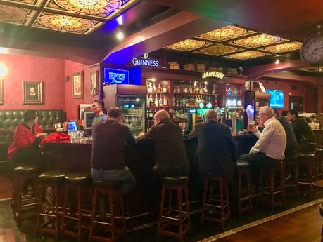 A cozy bar scene with people sitting on high stools at the bar. The bar is decorated with wooden details and neon signs, including one for Guinness, with various bottles of liquor on shelves behind the bar.