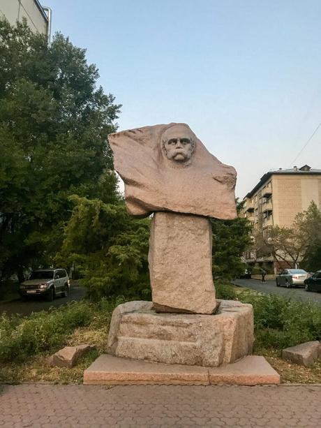 A statue of a man's head and shoulders carved into a rough stone block, standing outdoors in a small park surrounded by trees and a residential street.