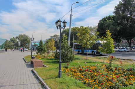 A city sidewalk lined with flowers and trees, next to a road with a bus driving by. The sky is clear and blue, with pedestrians walking along the path.