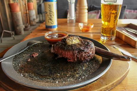 A juicy, seared steak served with sauce on a large plate, accompanied by a cold Heineken beer and a can of local beer in a wooden, rustic pub environment.