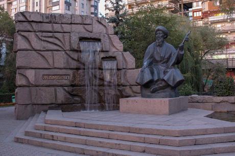 A bronze statue of a seated figure playing a traditional instrument next to a small waterfall, part of a monument inscribed with the name 