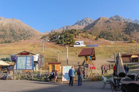 The Shymbulak ski resort area at the base of a mountain, featuring cable cars, signage, and a few people enjoying the sunny day, with the temperature showing +12°C.