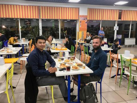 Alex Tiffany and a new Kazakh friend sit at a cafeteria table with a spread of traditional food, smiling at the camera. The cafeteria has colorful chairs and windows that look out to the city.