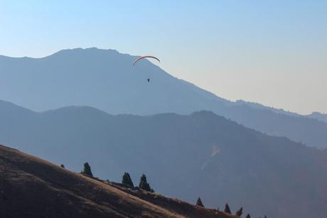 A person paragliding over a hilly landscape, with mountain ridges faintly visible in the background, creating a serene and open feeling.