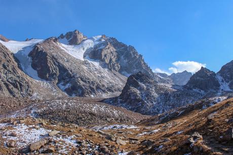 A rugged mountain landscape with snow-capped peaks and rocky slopes, under a bright blue sky. Patches of snow dot the ground in the foreground.