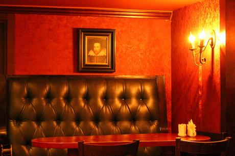 A warmly lit booth in a bar or pub, featuring a tufted black leather bench and a small wooden table. A portrait of a historical figure, possibly William Shakespeare, in a ruffled collar hangs on the deep red wall.