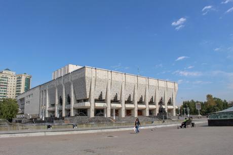 A large, brutalist-style concrete building with sharp, angular features. A few people are walking near the building, and a fountain sits in front.