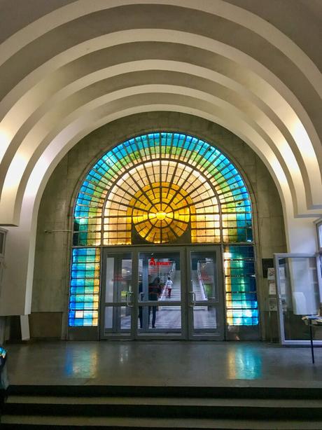 A stained-glass archway with geometric patterns of yellow, blue, and green over a set of double glass doors, inside the Arasan baths in Almaty.